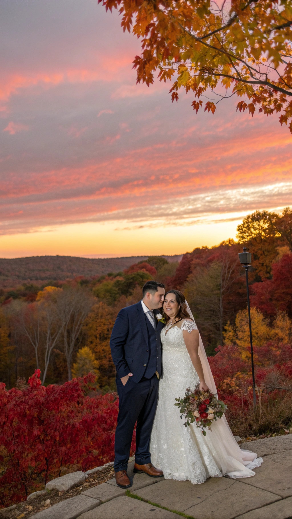 A couple in wedding attire poses against a stunning sunset with vibrant fall foliage.